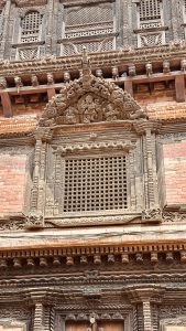 A close-up view of Bhaktapur museum intricately carved wooden architecture featuring a decorative window with a lattice design. Above the window, there are detailed carvings of figures and animals, framed by ornate borders. The surrounding walls display additional carvings and textures, showcasing traditional craftsmanship