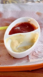 A white dipping dish with ketchup and mayonnaise sits on a napkin, with a single french fry on the side.