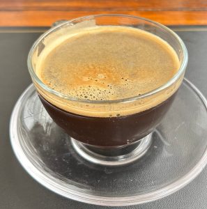 A glass cup of dark coffee sits on a small glass saucer.